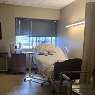A view of a patient room in a hospital, featuring a bed and natural light coming through the window.