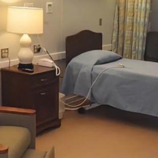 A hospital room featuring a bed with blue sheets, a nightstand with a lamp, and a curtain in the background.