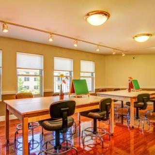 A well-lit classroom with several tables and chairs, featuring large windows and a polished wooden floor.