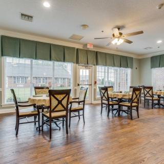 A spacious dining room with wooden flooring, featuring multiple tables and chairs arranged neatly, large windows that allow natural light to flood in, and ceiling fans for ventilation.