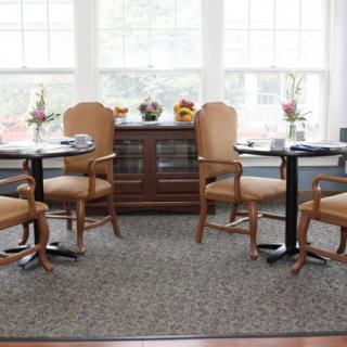 A cozy dining area with tables and chairs near large windows, adorned with flowers.