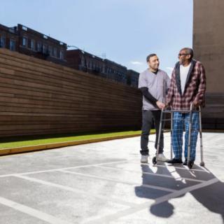 A caregiver assists an elderly man using a walker outdoors.