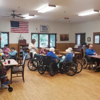 A community room in a nursing home where elderly residents are seated, some in wheelchairs, engaged in activities.