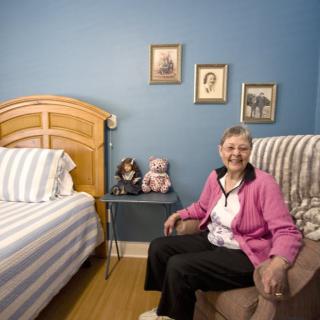 An elderly woman sitting comfortably on a chair in a cozy bedroom with blue walls and decorative items.