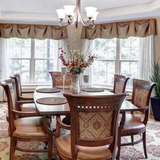 A cozy dining room with a wooden dining table and chairs, adorned with a chandelier and large windows with curtains.