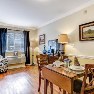 A cozy living and dining space featuring a wooden table with a complete table setting, stylish lamps, and wall art. The room is accented by wood flooring and natural light filtering through the window.