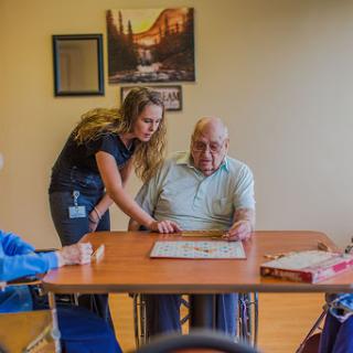 A caregiver assists an elderly man with a game while another resident looks on, promoting social interaction in an assisted living facility.