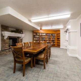 A cozy library room with a wooden table, chairs, bookshelves, and a fireplace.