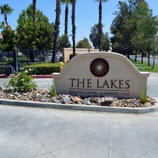 Entrance sign for 'The Lakes' community surrounded by palm trees and landscaped garden.