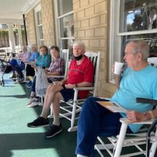 A group of seniors relaxing on a porch, sitting in rocking chairs during the day.