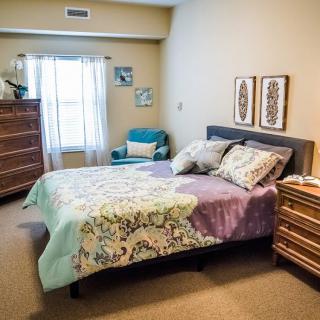 A cozy bedroom featuring a bed with patterned bedding, wooden dressers, and a comfortable chair by the window.