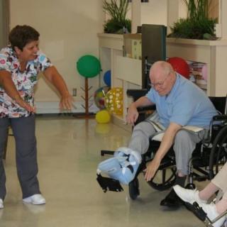 A caregiver assists an elderly person in a wheelchair with physical exercises in a nursing home setting.