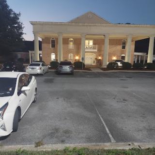 A view of a hotel with a well-lit entrance and parked cars in the lot during twilight.