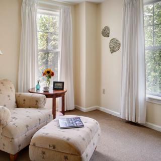 A cozy corner of a living room with a patterned armchair and ottoman, illuminated by natural light from large windows with curtains.