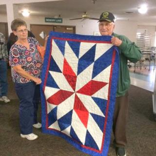 Two individuals proudly display a beautifully crafted quilt featuring a vibrant star design in red, white, and blue.