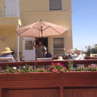 A group of people relax on a sunny patio under an umbrella, surrounded by flowers.