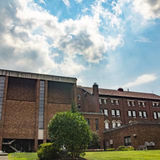 A brick building with a cloudy sky and lush greenery in the foreground.
