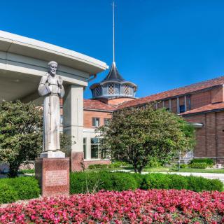 A statue stands in front of a brick building surrounded by vibrant flowers and greenery under a clear blue sky.