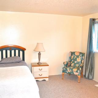 A simple bedroom with a bed, nightstand, lamp, and a patterned armchair near the window with curtains.
