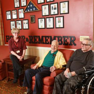 A group of elderly veterans sitting in a room adorned with military memorabilia and framed photos, symbolizing their service and dedication.