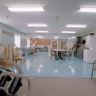 A spacious common area with seating and a kitchen in the background, featuring blue flooring and bright overhead lights.