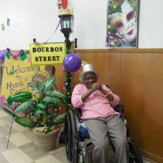 A festive Mardi Gras celebration with colorful decorations and a person in a wheelchair smiling in front of a Bourbon Street sign.