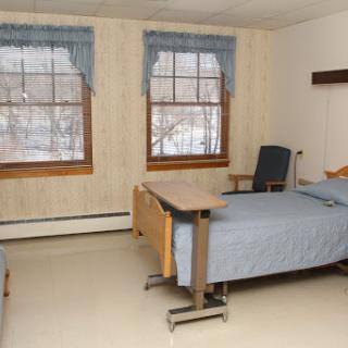 A simple and clean hospital room featuring a bed, a chair, and bright windows.