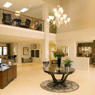 A spacious and elegant lobby with a chandelier, featuring a central table with flowers and a balcony above.