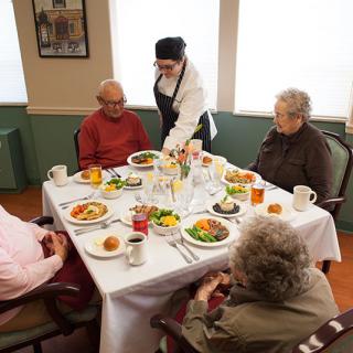 A group of seniors enjoying a meal together at a dining table with a server attending to them.