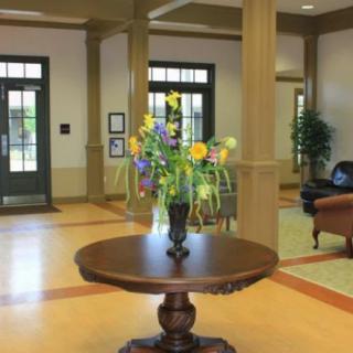 A lobby area featuring a central wooden table with a vase of colorful flowers, surrounded by comfortable seating and large windows.