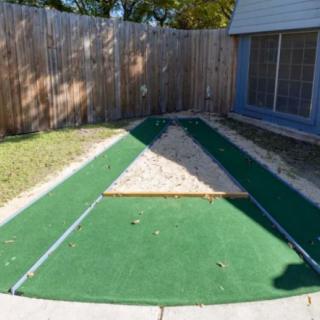 A small outdoor putting green with artificial turf and a sand trap, located in a fenced backyard.
