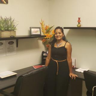 A woman stands in an office next to a desk with shelves and decorative plants.