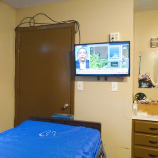 A simple room featuring a bed with a blue cover, a television mounted on the wall, and wooden furniture.