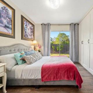 A cozy bedroom with a neatly made bed adorned with colorful pillows and a red blanket, complemented by artwork on the walls and a window with curtains letting in natural light.