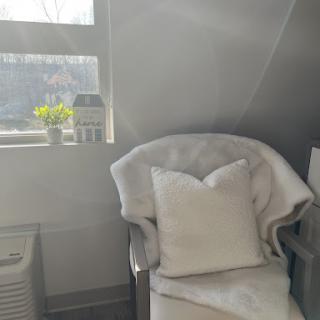 A cozy corner featuring a comfortable chair with a white pillow, bathed in natural light, complemented by a small plant on the windowsill.