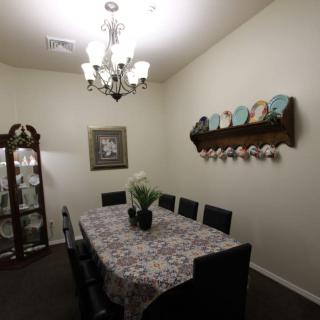A cozy dining room featuring a table with a floral tablecloth, surrounded by chairs, and illuminated by a chandelier. Decorative plates adorn the wall, and a vase with flowers sits in the center of the table.