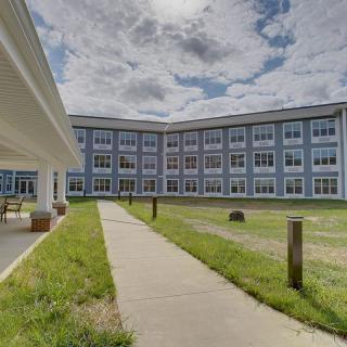 A modern building with multiple windows and a pathway leading through a grassy area under a cloudy sky.