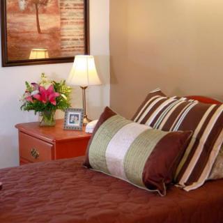 A cozy bedroom with a neatly made bed, striped pillows, and a nightstand featuring a lamp, flowers, and a framed photo.