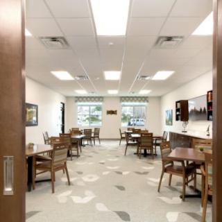 An interior view of a bright dining room with multiple tables and chairs, featuring large windows and ceiling lights.