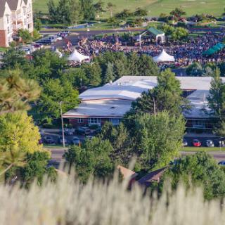 A scenic view of a large outdoor event with a gathering of people, surrounded by greenery and tents.