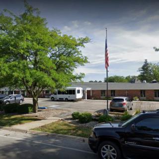 A parking area with several parked vehicles, surrounded by trees and a single-story building, featuring an American flag on a flagpole.