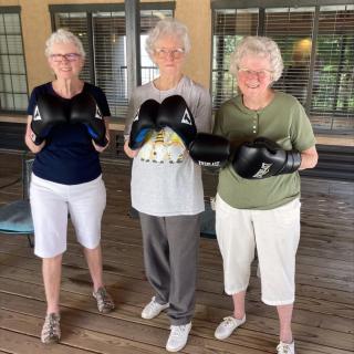 Three elderly women wearing boxing gloves stand together on a wooden deck.