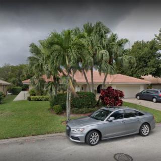 A suburban residential street with a house, palm trees, and parked cars.