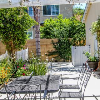 A sunny backyard patio with outdoor furniture surrounded by lush greenery and a wooden fence.