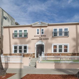 A two-story residential building with a stucco exterior and a small front yard behind a white fence.