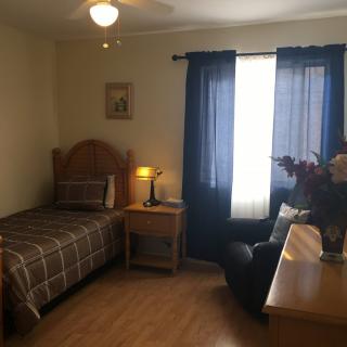 A cozy bedroom with a single bed, nightstand, and armchair next to a window with blue curtains.