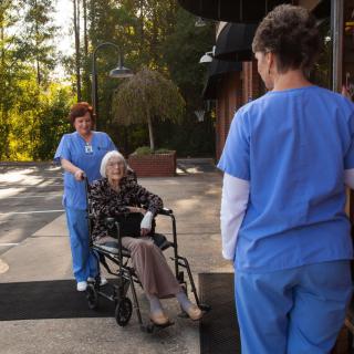 A nurse assists an elderly woman in a wheelchair outdoors, while another staff member looks on.