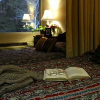 A cozy bedroom setting with a book and glasses on the bed, illuminated by warm lamplight.