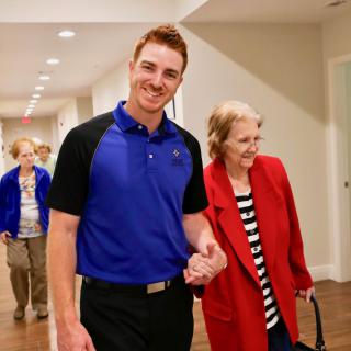 A caregiver walking with an elderly woman down a hallway, both smiling.