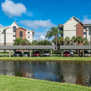 A view of residential apartments with palm trees and parked cars by a tranquil lake on a sunny day.